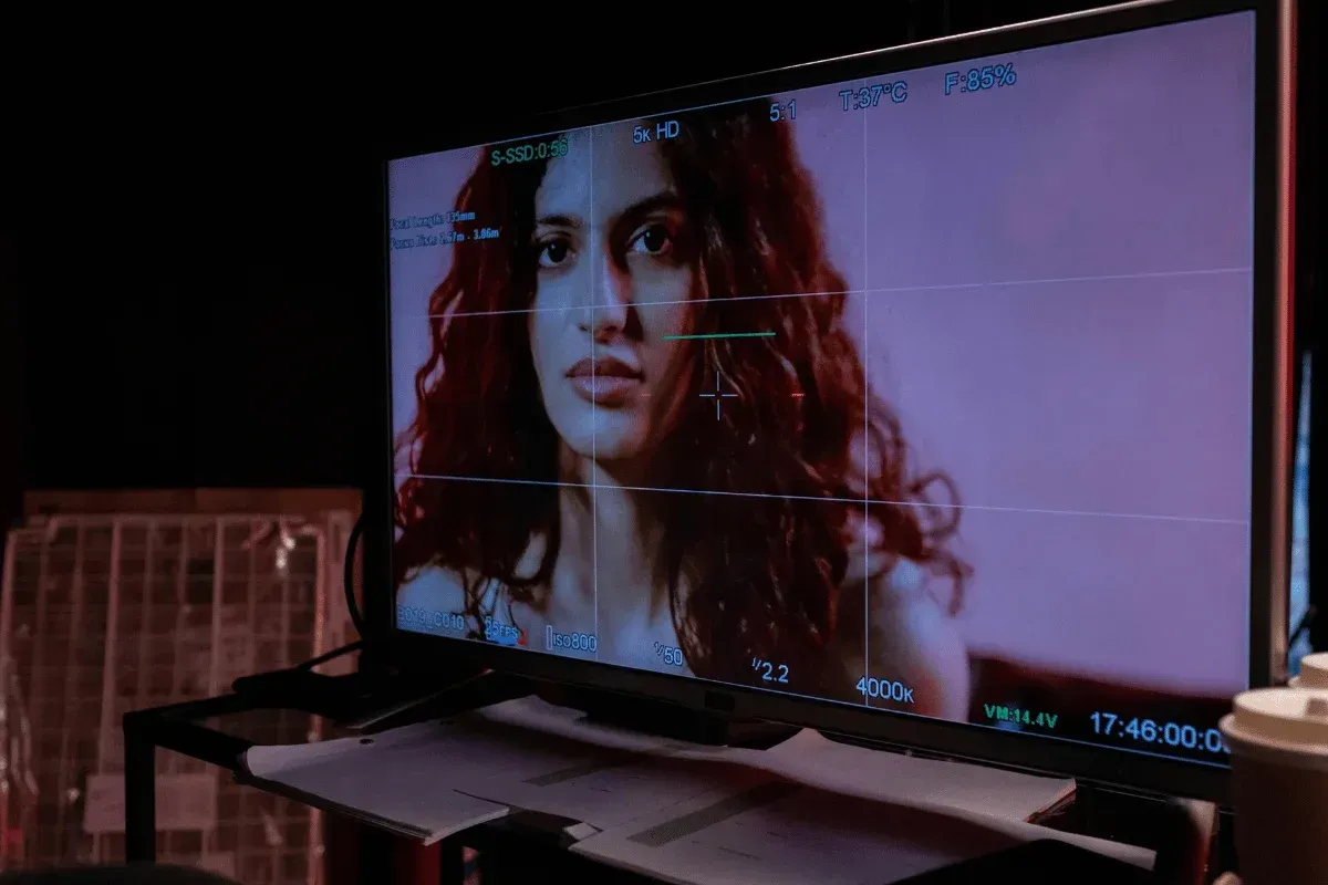 A monitor displays a scene with a person having long, curly red hair, set against a dimly lit background with technical settings visible.