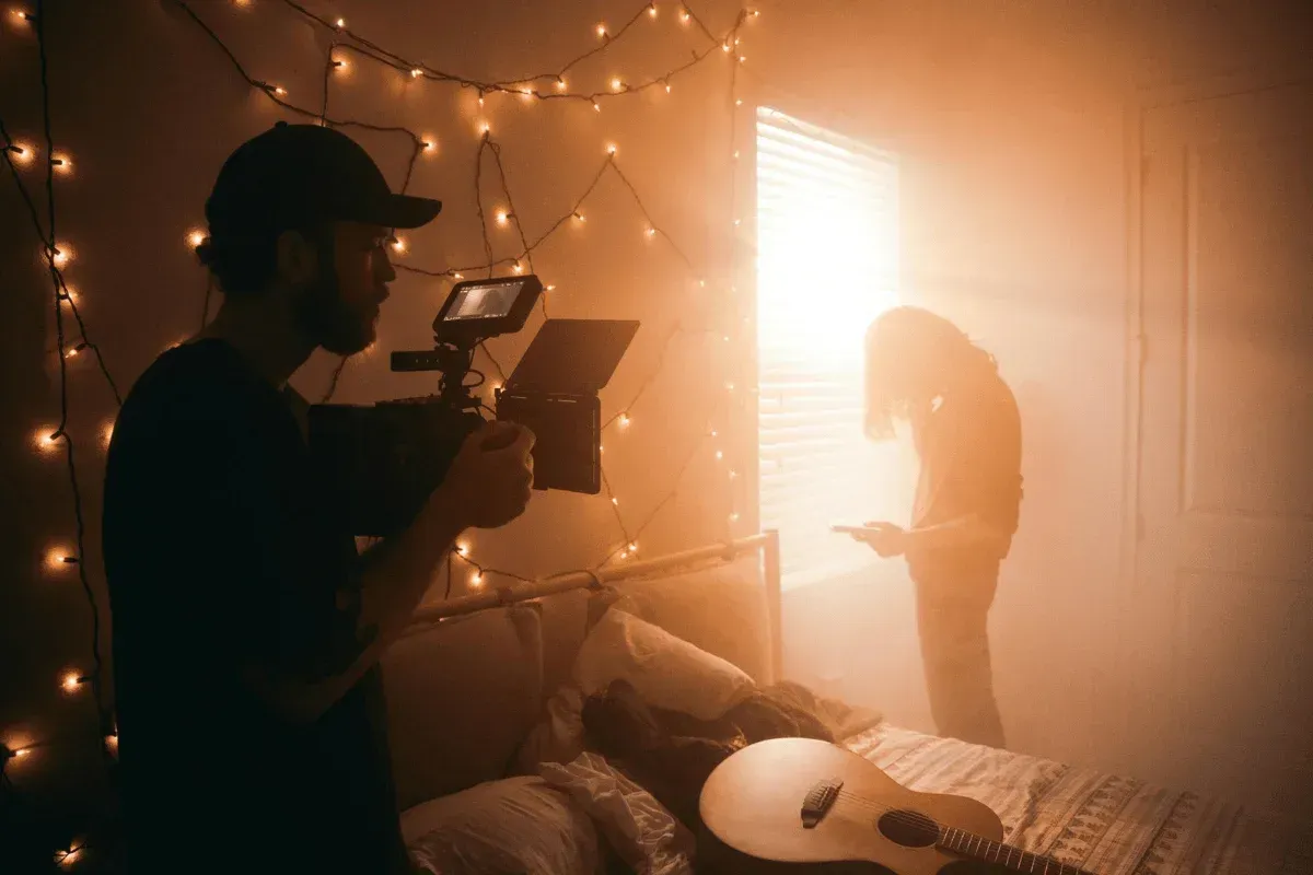Un cinéaste filme une personne près d’une fenêtre ensoleillée, dans une chambre chaleureuse éclairée par une guirlande. Une guitare posée sur le lit renforce l’atmosphère calme et artistique.