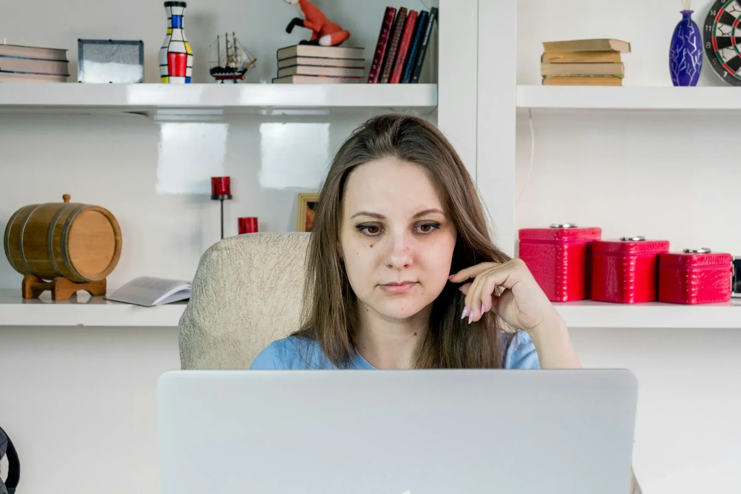 Une femme aux cheveux longs se concentre sur son ordinateur portable, assise devant une bibliothèque remplie de livres, un tonneau en bois et des objets décoratifs. L’atmosphère est chaleureuse et studieuse.