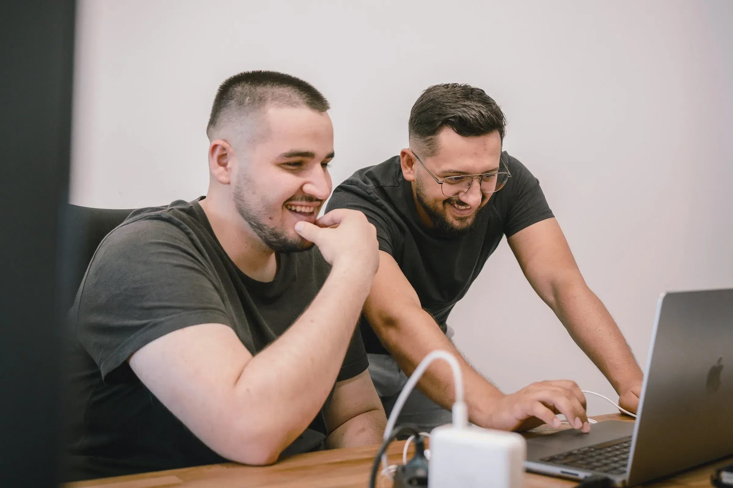 Deux hommes souriants travaillent sur un ordinateur portable à un bureau en bois, dans une atmosphère collaborative et positive. Ils portent des tee-shirts foncés décontractés.