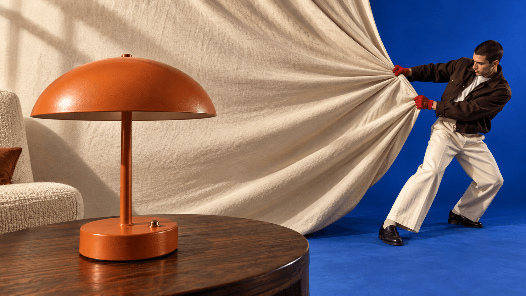 Orange mushroom table lamp on a walnut table as a stylist pulls back a cream backdrop to reveal a blue studio.