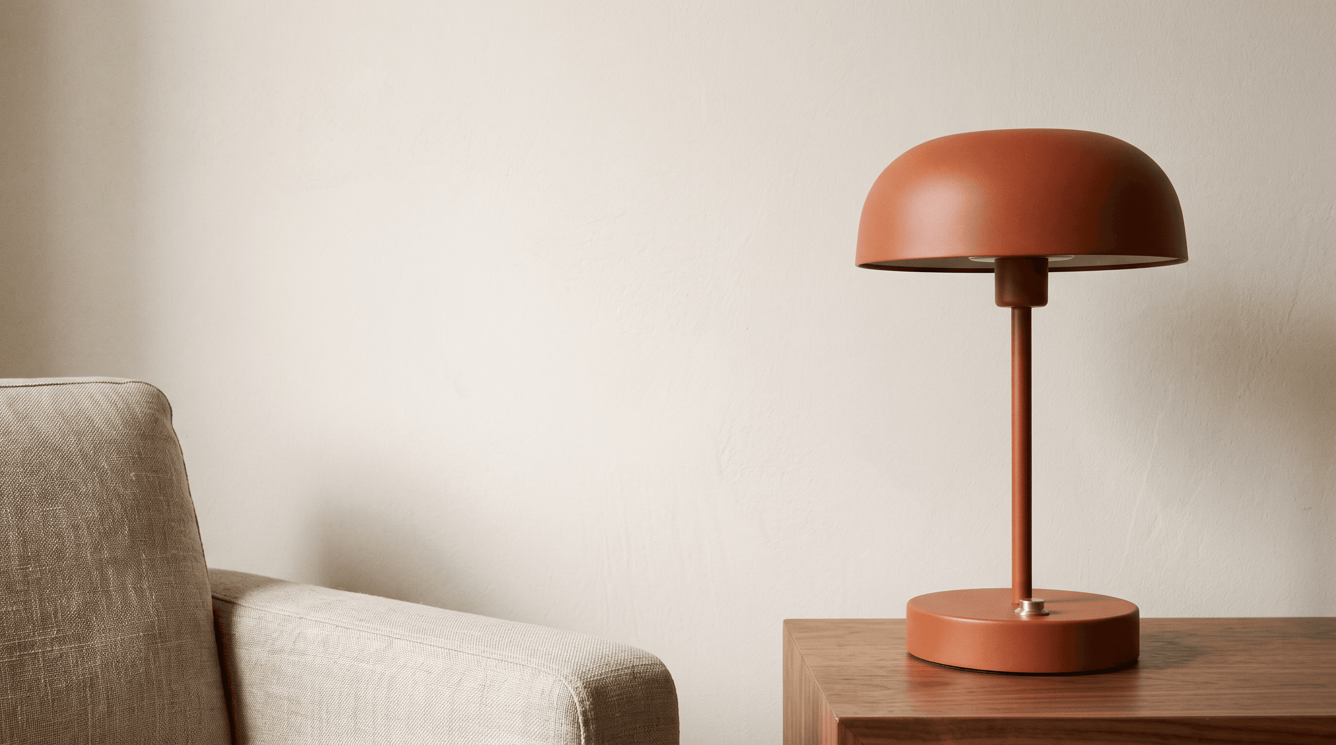 Orange table lamp on a walnut side table beside a linen armchair, with open cream wall space on the left.